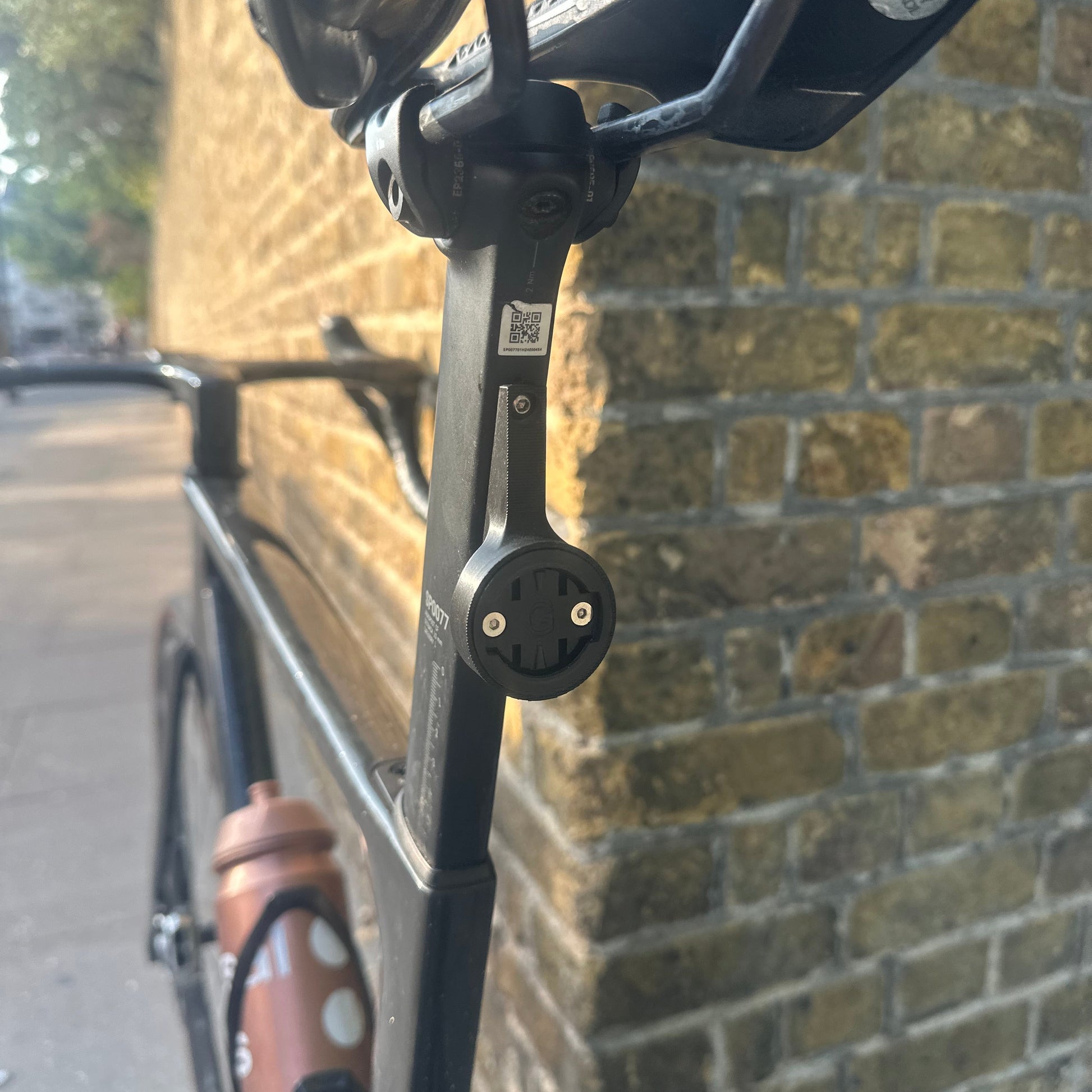 Close-up of a bicycle frame with a black mounting bracket attached near the handlebar area, photographed outdoors on a cobblestone surface.