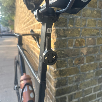 Close-up of a bicycle frame with a black mounting bracket attached near the handlebar area, photographed outdoors on a cobblestone surface.