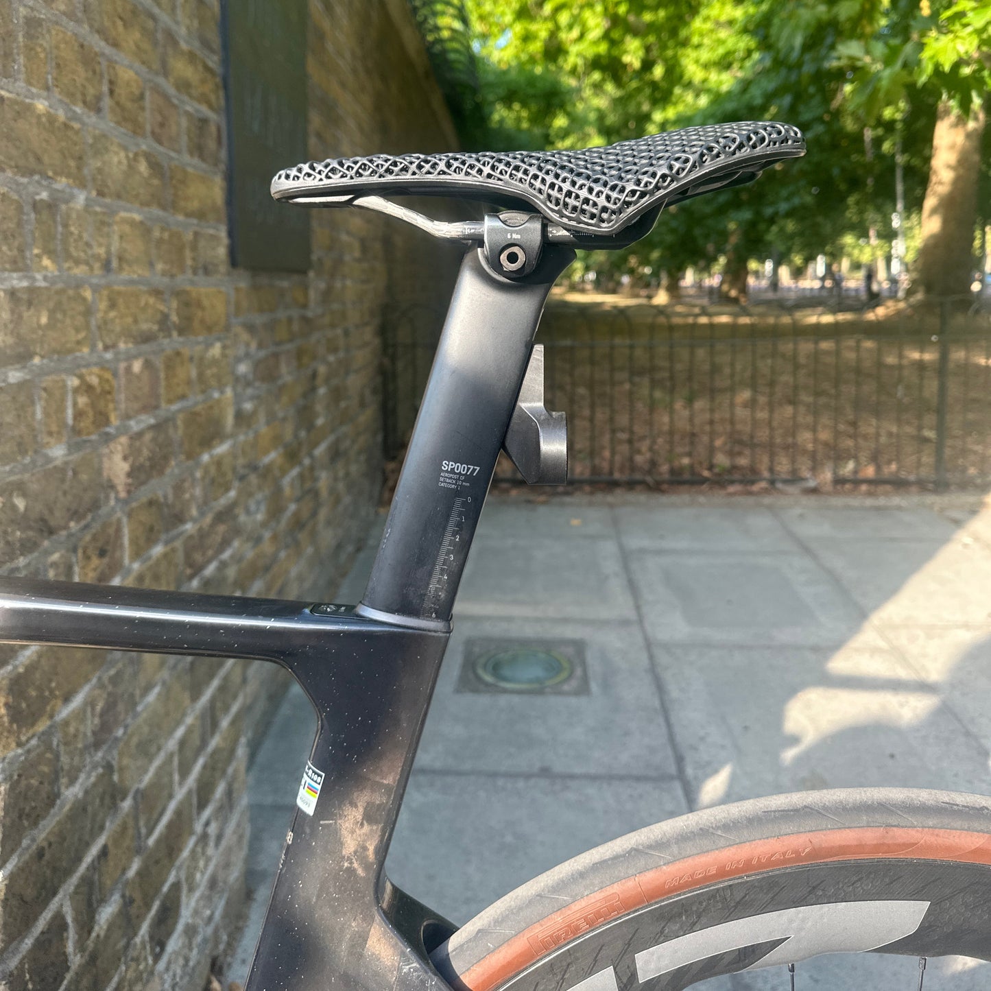 Close-up of a black road bicycle frame and saddle, photographed outdoors next to a brick wall with a paved sidewalk and green trees in the background.