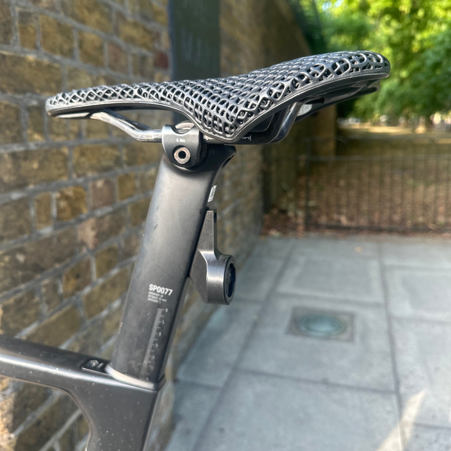 Close-up of a bicycle saddle mounted on a black seat post, photographed outdoors against a brick wall and paved path.