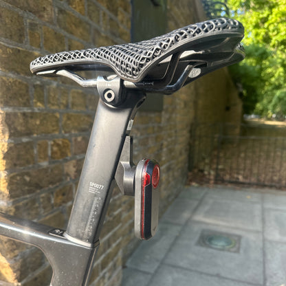 Close-up of a bicycle seat post with a black mesh saddle and a small rear reflector, photographed outdoors against a brick wall and paved path.