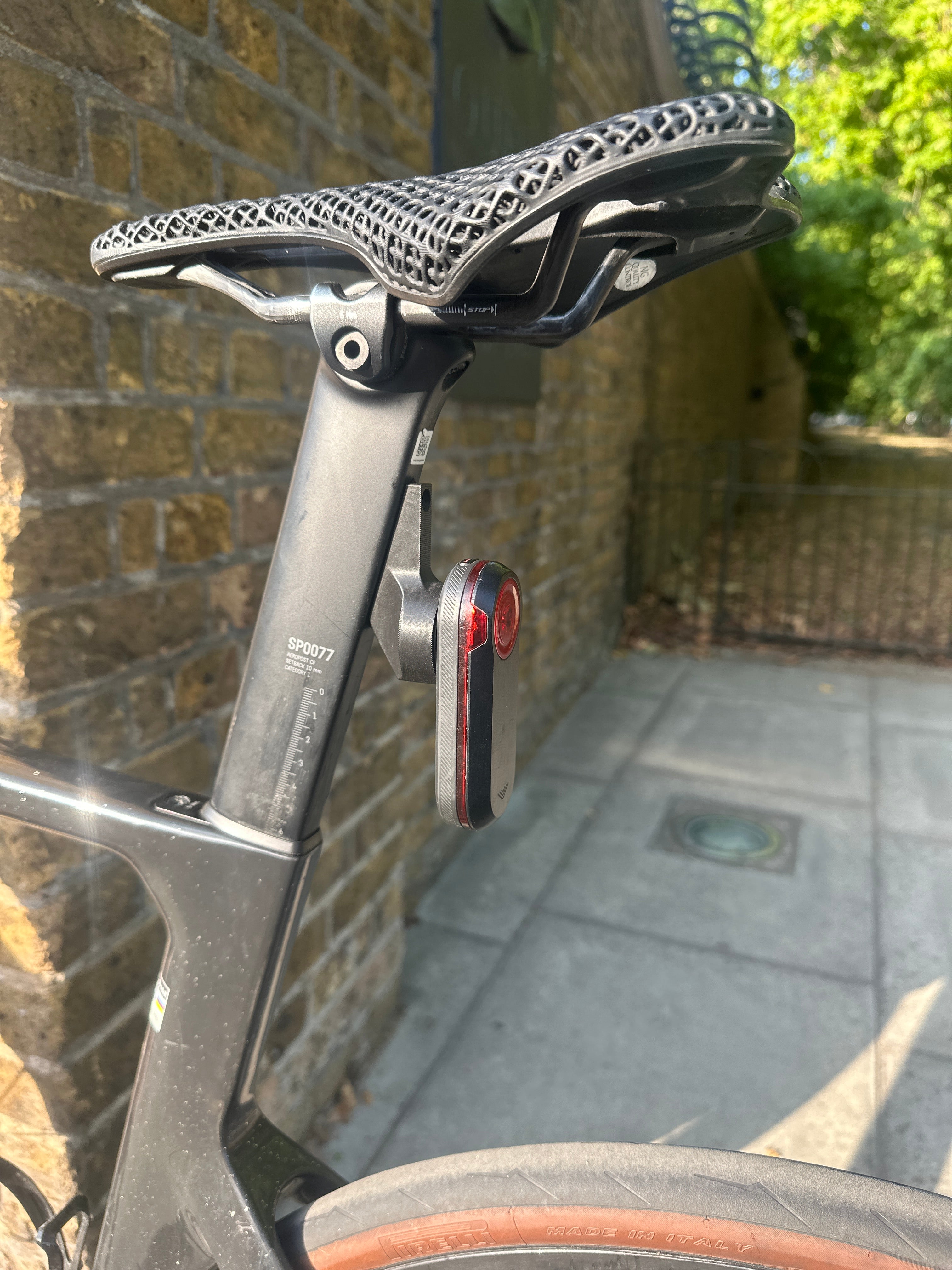Close-up of a bicycle seat and frame with a black mesh saddle and a small red-and-black rear reflector, photographed outdoors against a brick wall and paved path.