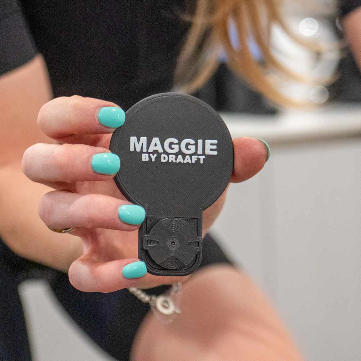 Black device with 'Maggie by Drafft' branding held by a person with teal nail polish.